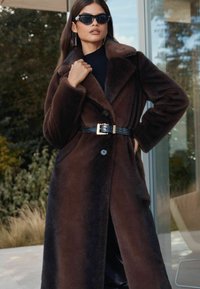 Barna műszőrme kabát, amelynek fűzött gallérja, dupla gombzárja van, és fekete krokodilmintás öv díszíti arany csattal.