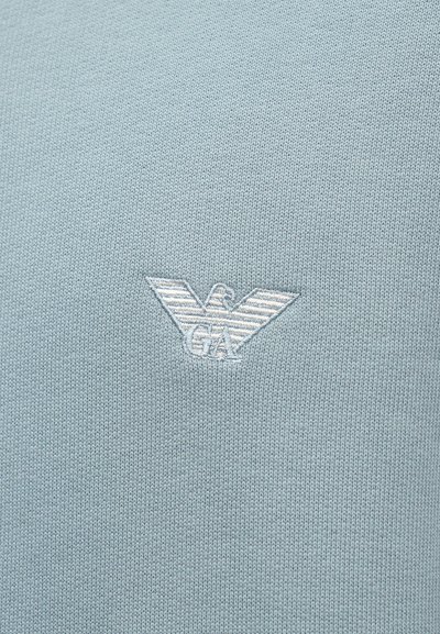 Ljusblå stickad tröja med en liten, broderad örnlogotyp i vitt på bröstet. Texturen är jämn och slät.