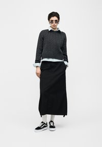 Άνθρωπος που φοράει στρογγυλά γυαλιά ηλίου, σκούρο γκρι πλεκτό πουλόβερ με πλεξούδες πάνω από ανοιχτό μπλε πουκάμισο, μαύρη μακριά φούστα, λευκές κάλτσες και μαύρα αθλητικά παπούτσια.