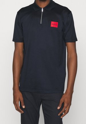 Navy polo shirt with a zippered collar, featuring a red rectangular logo patch with "HUGO" text. Smooth material and short sleeves.