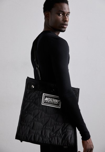 Borsa tote nera trapuntata con una patches logo Moschino, dotata di tracolla e superficie texturizzata. La borsa ha una forma squadrata e hardware in argento.