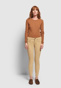 Mujer con pelo rojo que lleva un suéter marrón de canalé, pantalones ajustados beige y zapatos marrones tipo slip-on, de pie frente a un fondo gris claro.