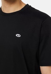 Černé bavlněné tričko s kulatým výstřihem. Má malé bílé oválné logo na hrudi. Krátké rukávy a hladký povrch.