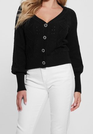 Femme portant un cardigan en maille noir boutonné avec motif torsadé et un pantalon blanc taille haute sur fond uni.