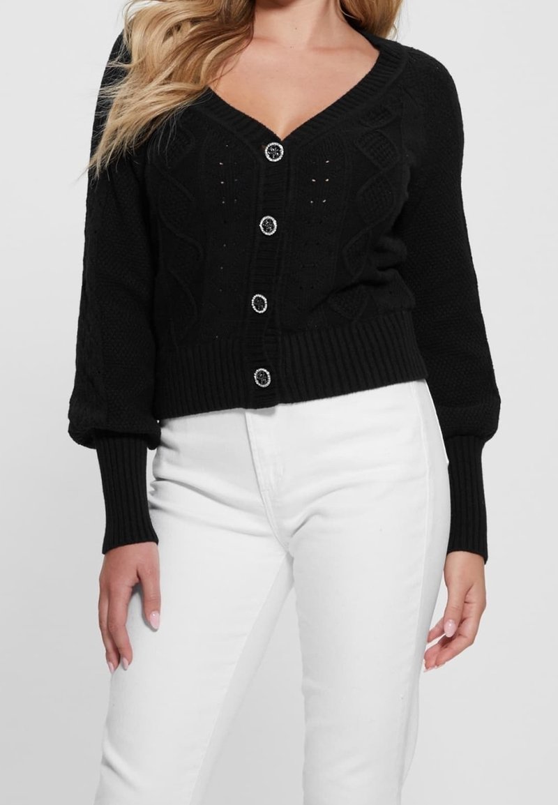 Femme portant un cardigan en maille noir boutonné avec motif torsadé et un pantalon blanc taille haute sur fond uni.