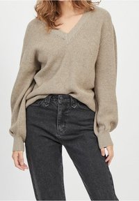 Pull en maille côtelée beige avec col en V, manches longues bouffantes, associé à un jean en denim noir taille haute. Texture douce et coupe ample.