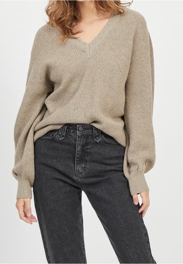 Pull en maille côtelée beige avec col en V, manches longues bouffantes, associé à un jean en denim noir taille haute. Texture douce et coupe ample.
