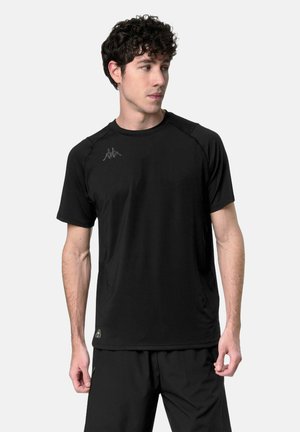 Uomo che indossa una maglietta sportiva nera a maniche corte e pantaloncini neri, che guarda a sinistra su uno sfondo chiaro e uniforme.