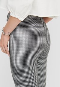 Zwart-witte houndstooth patroon leggings, nauwsluitend ontwerp met een hoge taille. Zichtbare hand rustend op de tailleband.