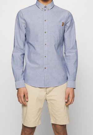 Chemise bleu clair à manches longues en coton, avec une poche poitrine, des boutons marron, et un ourlet courbé.