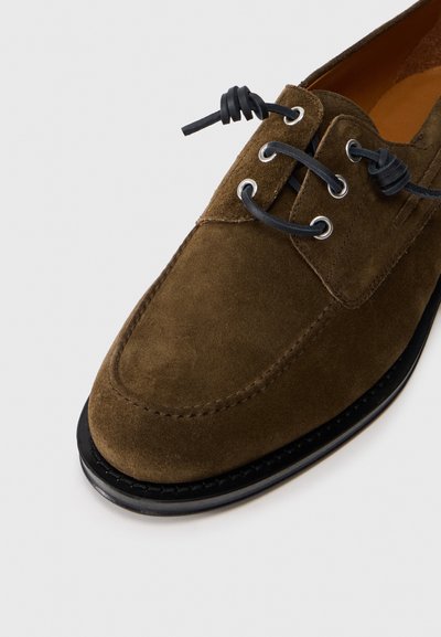 Chaussure en daim brun avec un bout arrondi, lacets noirs à travers des œillets en métal et une semelle en caoutchouc noir contrastante. Détails de surface texturés visibles.