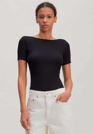 Vrouw draagt een aansluitende zwarte top met korte mouwen en witte broek met hoge taille, met één hand in de zak, tegen een effen achtergrond.