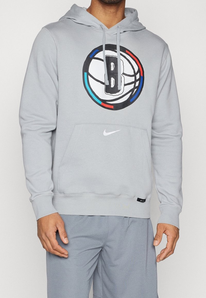 Homme portant un sweat à capuche gris clair avec le logo de basket-ball des Brooklyn Nets et un swoosh Nike blanc, associé à un short gris assorti.