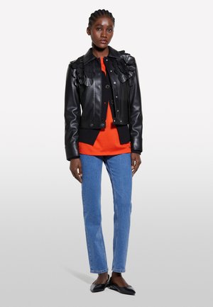 Veste en cuir noire avec des accents floraux texturés, associée à un haut orange, un jean bleu clair et des ballerines noires, sur fond blanc.