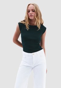 Top senza maniche di un verde scuro con texture a coste, abbinato a jeans bianchi a vita alta con chiusura frontale a bottone e tasche.