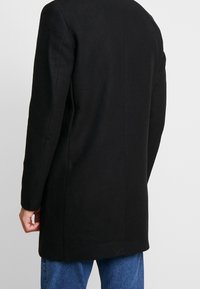 Cappotto di lana nero con maniche lunghe, vestibilità sartoriale e taglio dritto. Design minimalista senza hardware o accenti visibili.