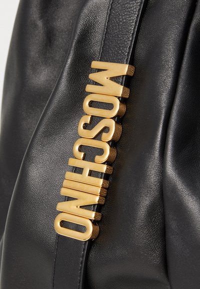 Juodas odinis krepšys su didelėmis auksinėmis raidėmis, rašančiomis "MOSCHINO", prisegtomis vertikaliai prie diržo.
