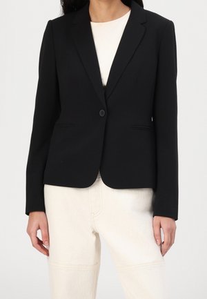 Frau trägt einen taillierten schwarzen Blazer mit einem Knopf über einem weißen Top und hellen Hosen, steht vor einem schlichten Hintergrund.