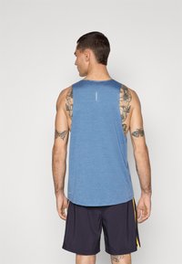 Blå atletisk tanktop lavet af letvægtsstof, med rund halsudskæring og rygdesign, parret med mørke shorts og synlige tatoveringer.