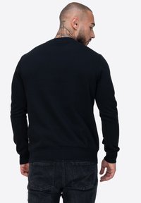 Sudadera negra con cuello redondo, mangas largas, puños y bajo acanalados. La tela es suave con una textura lisa, sin patrones visibles.