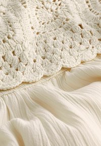 Crochet en dentelle dans un tissu léger crème, featuring des points texturés et des détails froncés pour une apparence fluide et superposée.