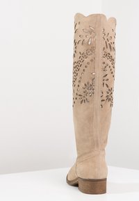 Beżowe zamszowe kozakami do kolan z wyciętymi wzorami kwiatowymi, falbaniastym wykończeniem na górze oraz niskim brązowym obcasem. Gładka tekstura i dopasowany krój.