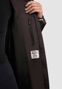 Mustan toppatakin sisäpuoli, jossa vetoketjullinen tasku ja etiketti, jossa lukee "Lämmin halaus kankaasta." Sileä tekstuuri, kevyt materiaali.