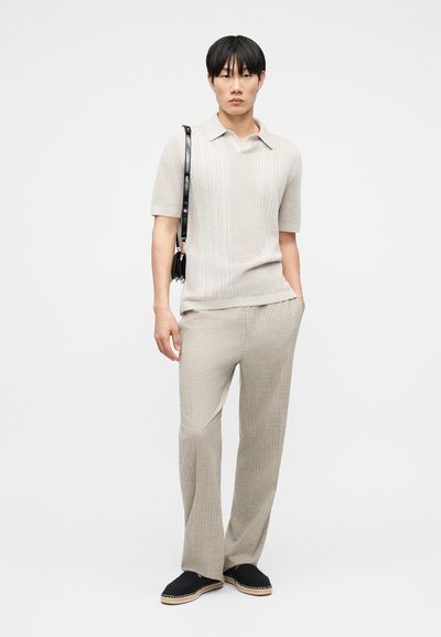 Νεαρός άνδρας που φοράει μπεζ υφασμένο πόλο με κοντά μανίκια και αντίστοιχο παντελόνι, με μαύρα εσπαντρίγιες και μαύρη τσάντα ώμου.