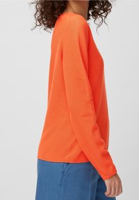 Pull en maille texturée orange avec manches longues et col rond, associé à un denim bleu. Le design est décontracté et ajustement ample.