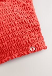 Korallszínű párna texturált, gyűrt szövettel és hullámos szegéllyel. Egyik sarkában egy kis, ezüst logó díszíti.