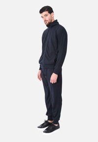 Giacca a zip navy con collo alto, abbinata a pantaloni jogger coordinati e sneakers nere. Tessuto morbido, vestibilità comoda e tasche laterali.