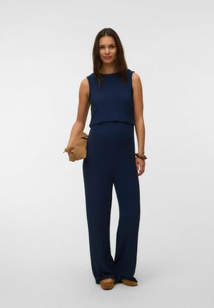 Kvinde iført en mørkeblå ærmeløs jumpsuit, der holder en lysebrun clutch, med matchende lysebrune sko og et brunt armbånd på sin højre håndled.