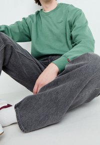 Sudadera verde con cuello redondo y logotipo bordado, combinada con jeans de corte relajado en gris oscuro y zapatillas blancas con detalles en burdeos.