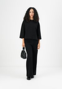 Schwarzes, strukturiertes Crop-Top mit weiten Ärmeln, kombiniert mit weit geschnittenen schwarzen Hosen. Hält eine kleine schwarze Handtasche. Glattes Material und Silhouette.