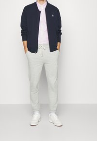 Námořnická bombardovací bunda, růžová pruhovaná košile, šedé tepláky a bílé tenisky. Bunda má logo a žebrované manžety; tepláky mají stahovací pas.