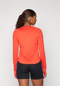 Langarmshirt in leuchtendem Orange mit einem runden Ausschnitt und Rückendetail mit weißen Streifen. Kombiniert mit schwarzen Shorts.