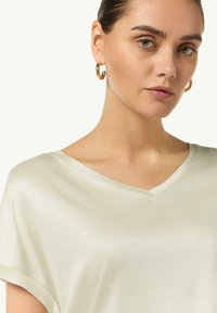 Jasny, t-shirt z dekoltem w serek, wykonany z gładkiego materiału, z krótkimi rękawami i luźnym krojem. Modelka nosi złote kolczyki w kształcie kół.