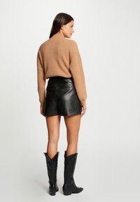 Pull en maille marron, design col montant, associé à un short en cuir noir et des bottes de cowboy noires avec des coutures détaillées et un bout pointu.