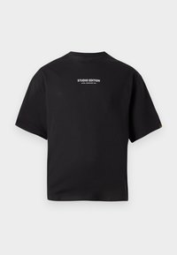 Juoda trumpomis rankovėmis mar�škinėliai su balta užrašu "STUDIO EDITION ALPHA INDUSTRIES INC." centre ant krūtinės prieš paprastą foną.