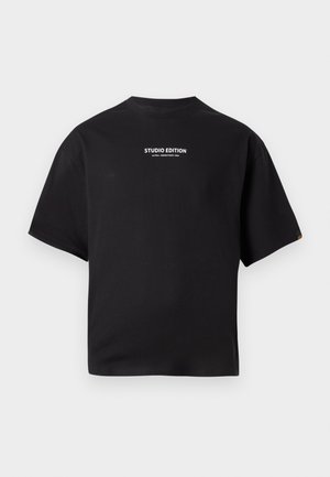 Černé tričko s krátkým rukávem s bílým textem "STUDIO EDITION ALPHA INDUSTRIES INC." umístěným uprostřed na hrudi na jednobarevném pozadí.