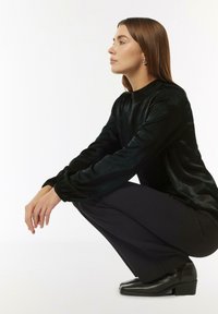 Schwarzes Samtoberteil mit langen Ärmeln, stehendem Kragen und gerafften Bündchen, kombiniert mit schwarzen Hosen und ankle Boots mit geometrischem Design.