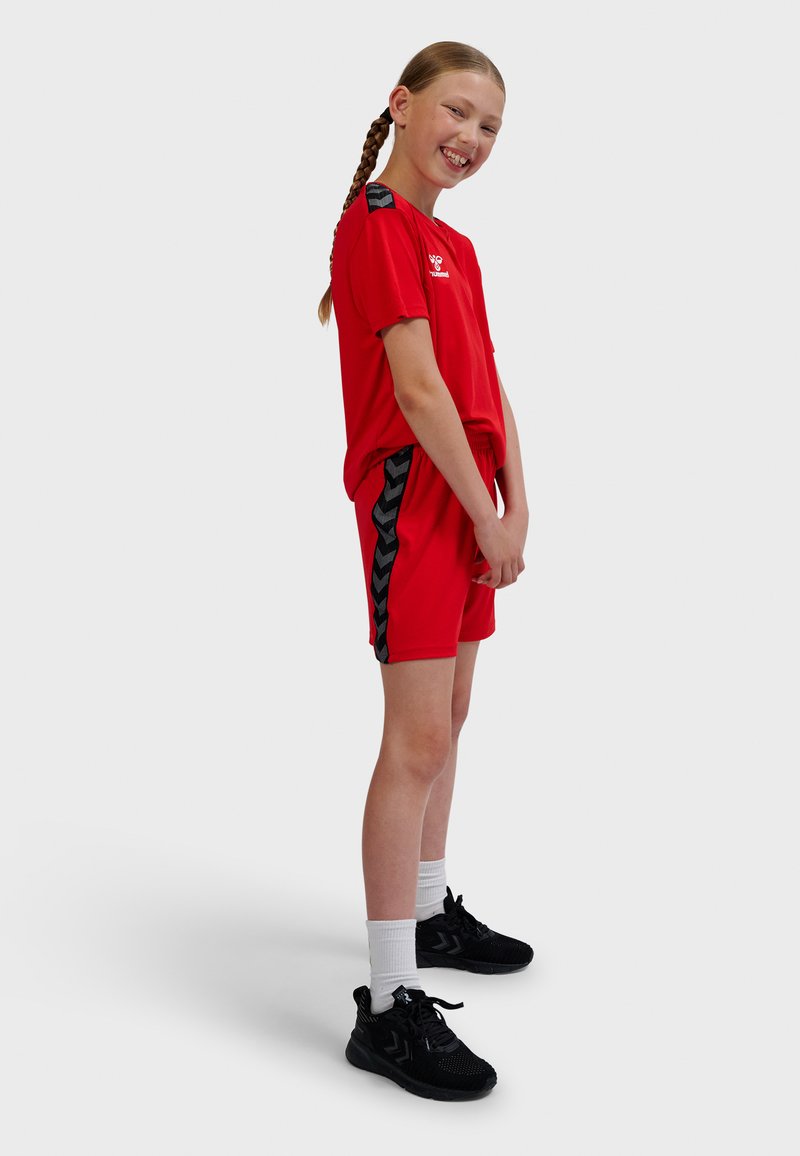 Rode sport T-shirt en korte broek set, met een zwart chevronpatroon op de zijnaden van de korte broek. Zwarte sneakers en witte sokken.