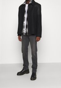 Svart jacka med läderärmar, över en vit och svart rutig skjorta. Grå jeans och svarta ankelskor fulländar outfiten.