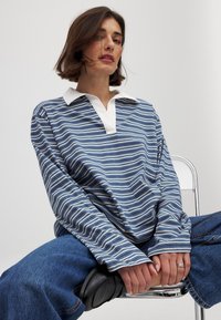 Camicia a maniche lunghe a righe blu e bianche, con colletto bianco e vestibilità morbida. Il modello indossa jeans scuri e scarpe nere.