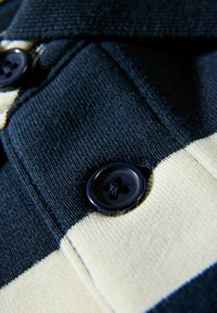 Granatowo-kremowa koszulka polo w paski z fakturowanej tkaniny, kontrastujących kolorów oraz czarnym guzikiem z czterema dziurkami.