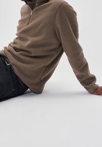 Pullover in pile marrone con colletto a mezze zip, superficie texturizzata e polsini a coste, abbinato a jeans neri e una cintura visibile.