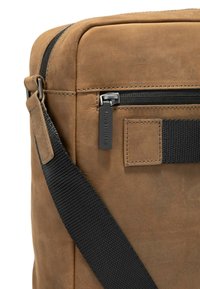 Braune Lederhandtasche mit glatter Textur, schwarzem Reißverschluss und verstellbarem schwarzen Gurt. Mit einem kleinen Anhänger mit "Strellson"-Markierung.