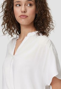 Blouse blanche avec une coupe décontractée, manches courtes et un petit col montant. Texture lisse, patte de boutonnage à l'avant avec un décolleté en V subtil.