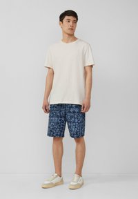 Licht beige katoenen t-shirt gecombineerd met blauwe patroonshorts met bloemendessins. Model draagt witte sneakers met beige accenten.