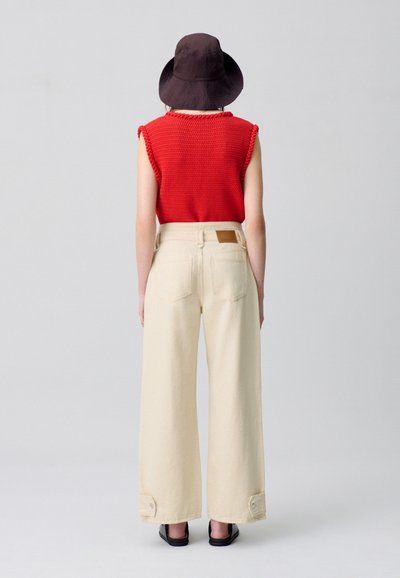 Top sin mangas rojo de punto, pantalones amplios crema con puños abotonados y un sombrero tipo cubo marrón. Diseño simple, tejido ligero, colores neutros.